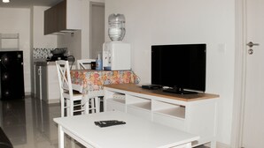 A compact kitchen with a refrigerator, microwave, and water dispenser. A dining table with chairs is positioned in front of a television. - Nice And Spacious 3Br At Meikarta Apartment (Cikarang)