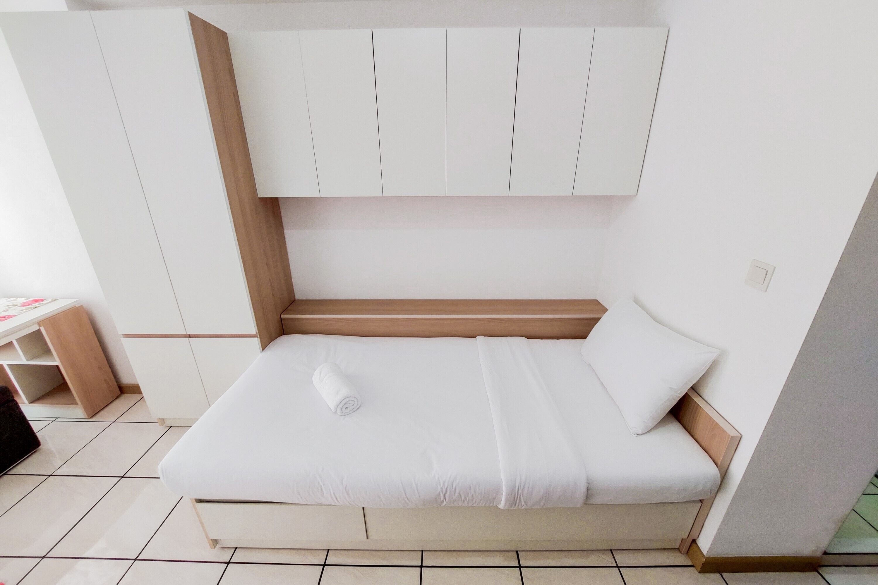 Una cama bien hecha con colchón y almohadas blancas, cabecera de madera y un armario blanco encima.