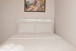 A neatly made bed with white bedding and pillows, a plain wall, and a framed painting above the bed.