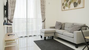 A modern living room with a grey sofa, a small table, and a large window with sheer curtains. - Comfrot 2Br At Sky House Bsd Apartment (Pagedangan)