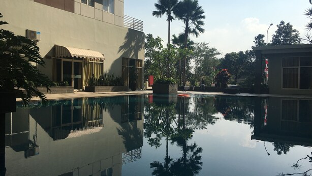 A swimming pool with clear reflections, surrounded by buildings and palm trees. - Cozy 2Br At Pinewood Apartment Jatinangor (Cileunyi)