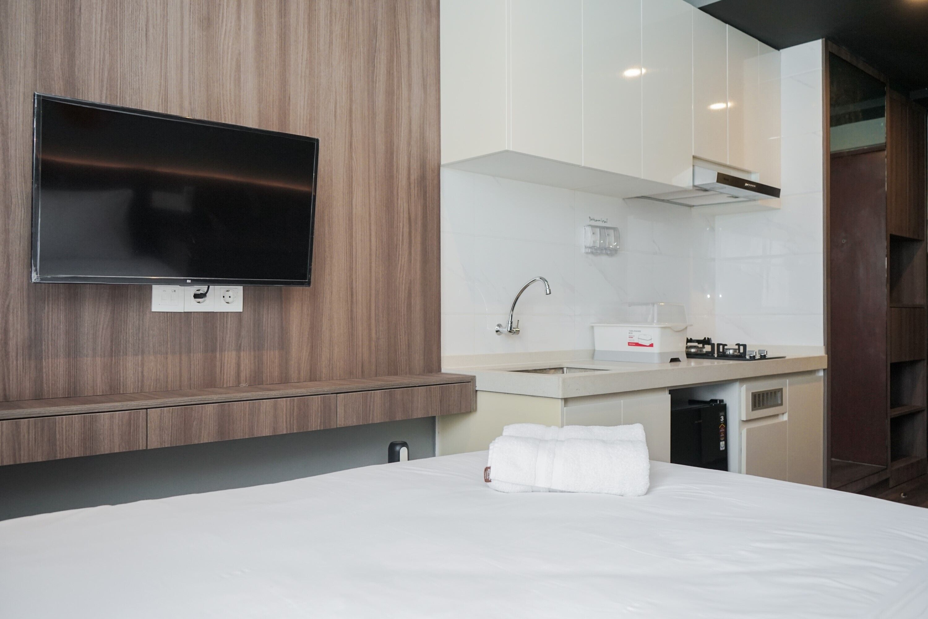 A modern hotel room with a flat-screen TV mounted on a wooden wall, a kitchenette with a sink and stove, and a neatly made bed with a white towel.