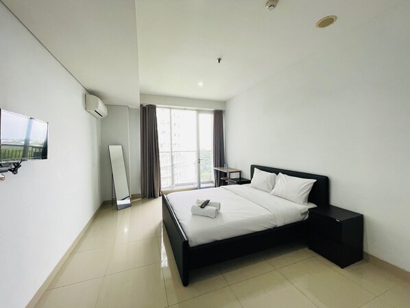 A modern hotel room with a large bed, a flat-screen TV mounted on the wall, a small table, and a window with curtains.