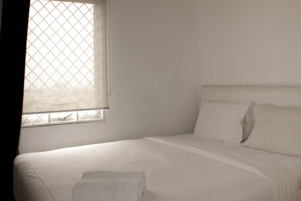 Ein Einzelbett mit weißer Bettwäsche, ein Fenster mit Jalousien und eine schlichte Wand.