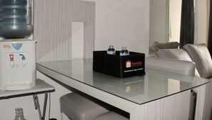 A hotel room with a glass-top table, a water dispenser, a black box with water bottles, a sofa, and a window with curtains. - Comfort 2Br Apartment At Mediterania Palace Residence (Jakarta)