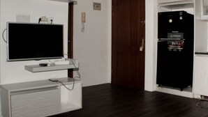 A room with a flat-screen TV mounted on a white shelf, a black refrigerator with a freezer compartment, and a wooden door. - Comfort 2Br Apartment At Mediterania Palace Residence (Jakarta)