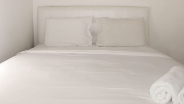 A neatly made bed with white linens and pillows, a rolled towel on the side, and a plain wall. - Comfort 2Br Apartment At Mediterania Palace Residence (Jakarta)