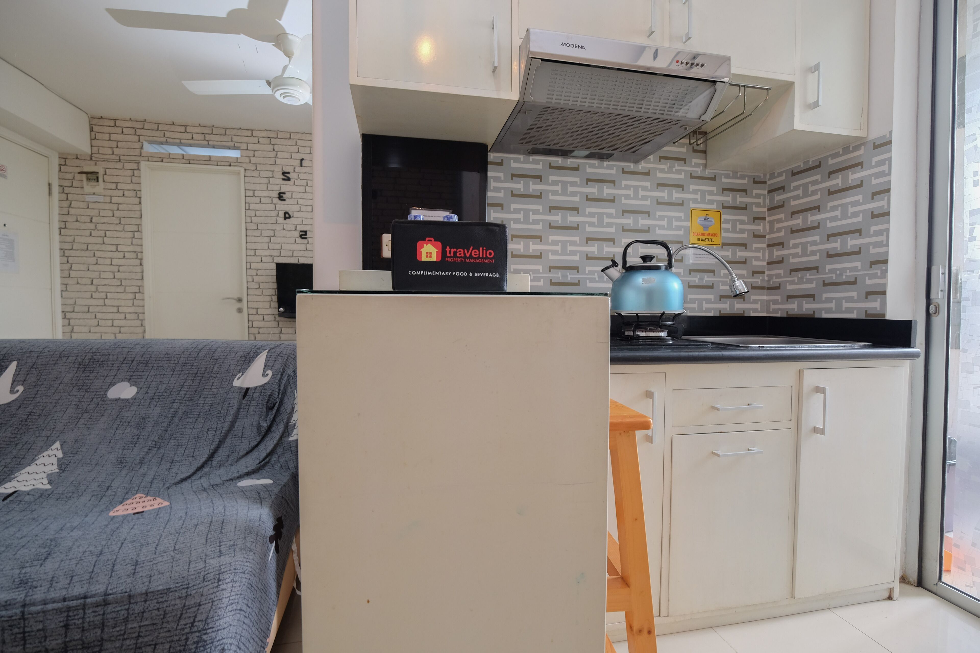 A compact kitchen with a microwave, kettle, and a travelio sign, a sofa with a blanket, and a door with a window.