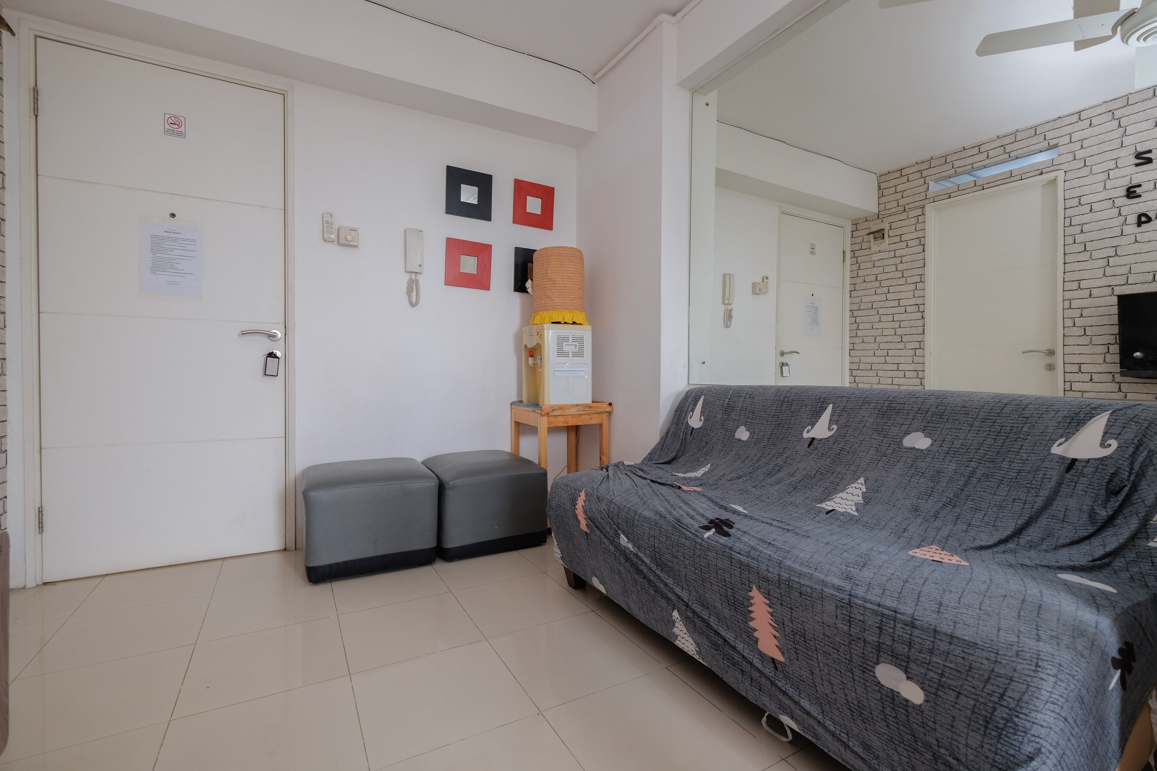 A room with a grey sofa, two grey ottomans, a small wooden table with a lamp, a brick-patterned wall, and a white door with a sign.