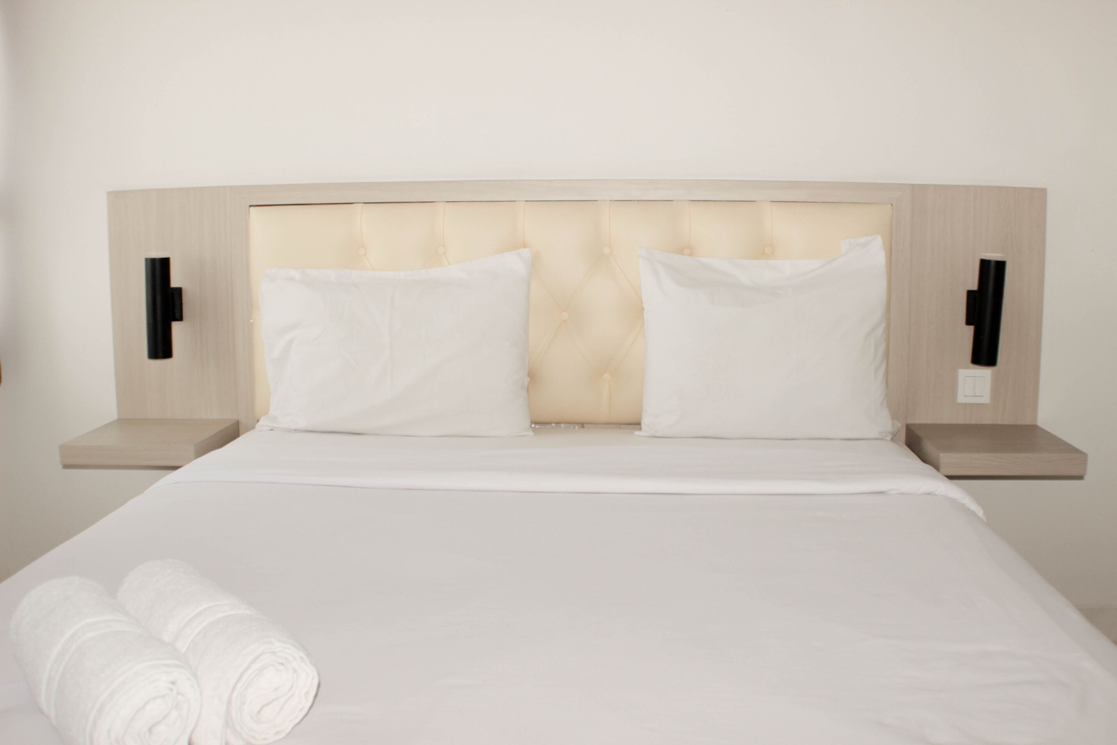 Un lit bien fait, avec du linge de lit blanc et deux oreillers, une tête de lit en bois et des tables de chevet avec des lampes noires.
