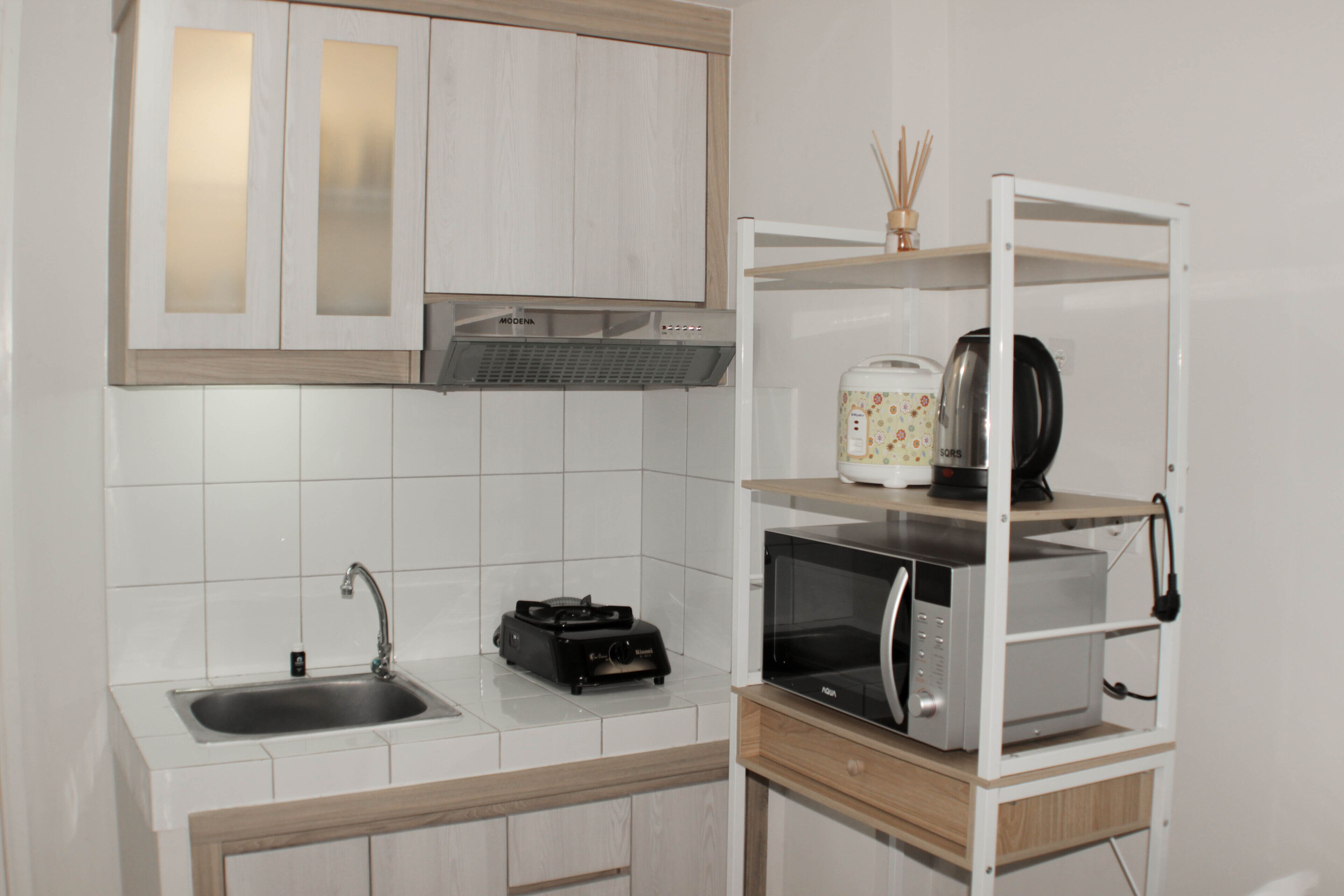 Una cocina con armarios blancos, un lavamanos de acero inoxidable, un microondas y una cafetera sobre una estantería blanca.