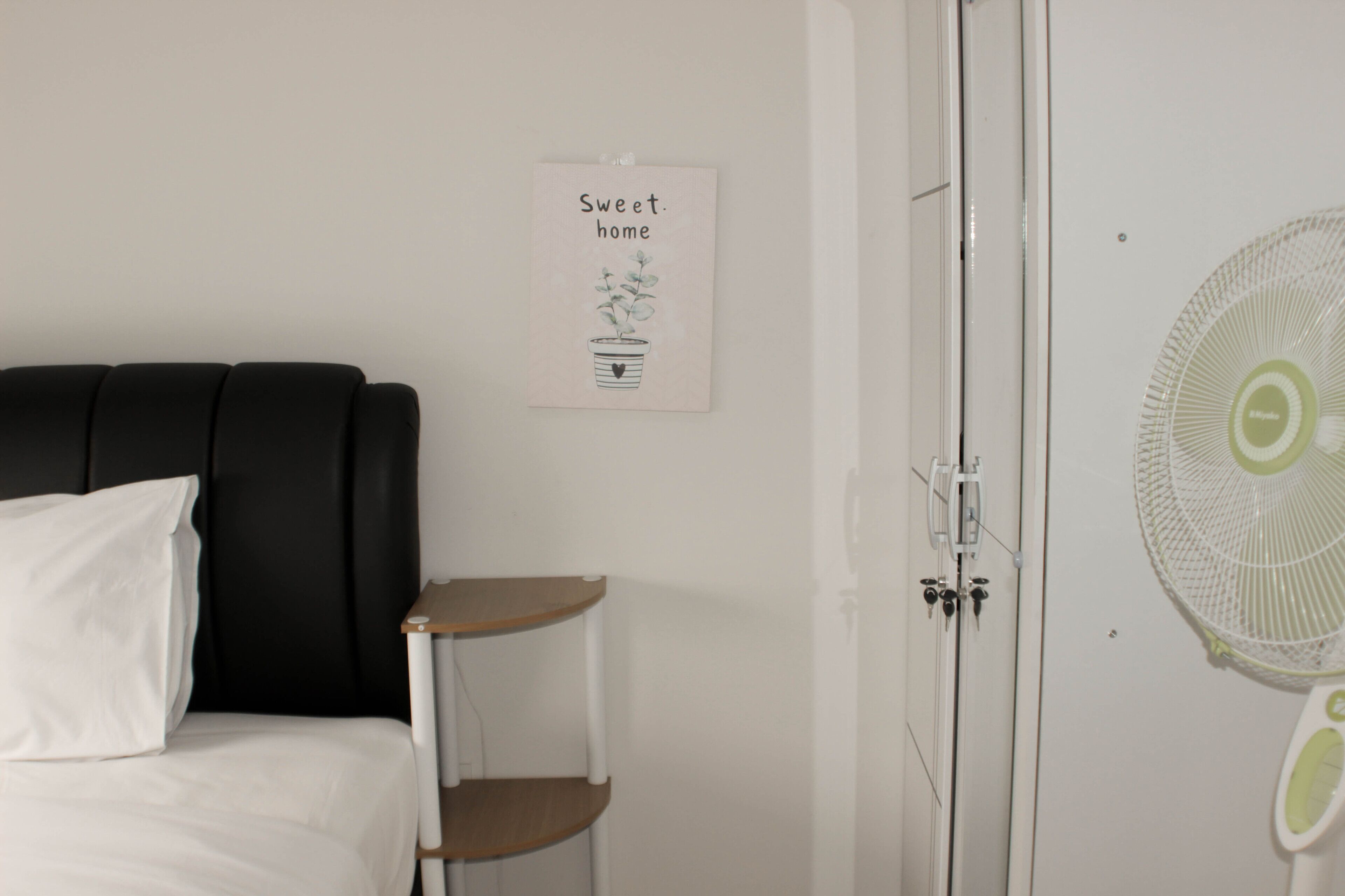 Ein Schlafzimmer mit einem Bett, einem Nachttisch, einem Wandventilator und einem Poster an der Wand.