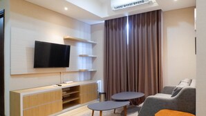 A modern living room with a flat-screen TV mounted on the wall, a wooden entertainment unit, two grey sofas, and a small round table. - 3Br Luxurious And Elegant Apartment At Grand Sungkono Lagoon (Surabaya)