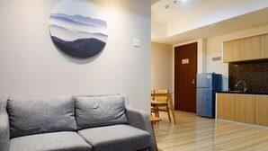 A modern living room with a grey sofa, a round wall art piece, a small table, and a kitchen area with wooden cabinets and a refrigerator. - 3Br Luxurious And Elegant Apartment At Grand Sungkono Lagoon (Surabaya)