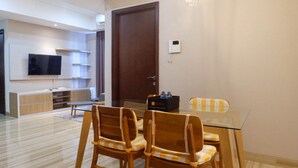 A modern dining area with a glass table and wooden chairs, a television on the wall, and a large door. - 3Br Luxurious And Elegant Apartment At Grand Sungkono Lagoon (Surabaya)