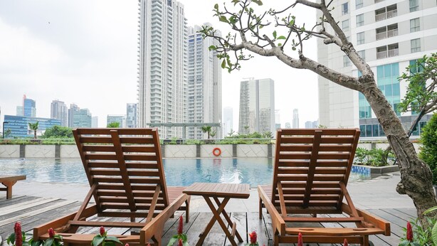A poolside area with wooden lounge chairs and a table, surrounded by greenery and a view of high-rise buildings.