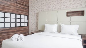 A neatly made bed with white linens and two rolled towels, a headboard with a wooden panel design, and a patterned wallpaper with a leaf motif.