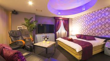 A hotel room with a bed, a massage chair, a TV, a minibar, and a small table with a bottle and glasses.