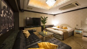 A modern hotel room with a black leather sofa, a bed with a white headboard, a small table with a plant, a TV, and a minibar.