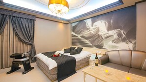 A modern hotel room with a bed, a sofa, a massage chair, a table with candles, and a large abstract painting on the wall.
