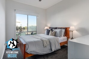 A bedroom with a large window, a bed with grey bedding, two bedside lamps, and a white bedside table.