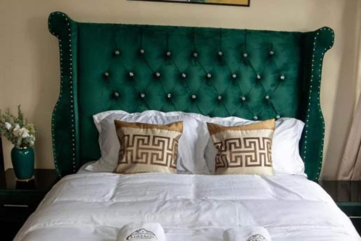 A bed with a green upholstered headboard, white bedding with patterned pillows, and a decorative pillow with a Greek key design.