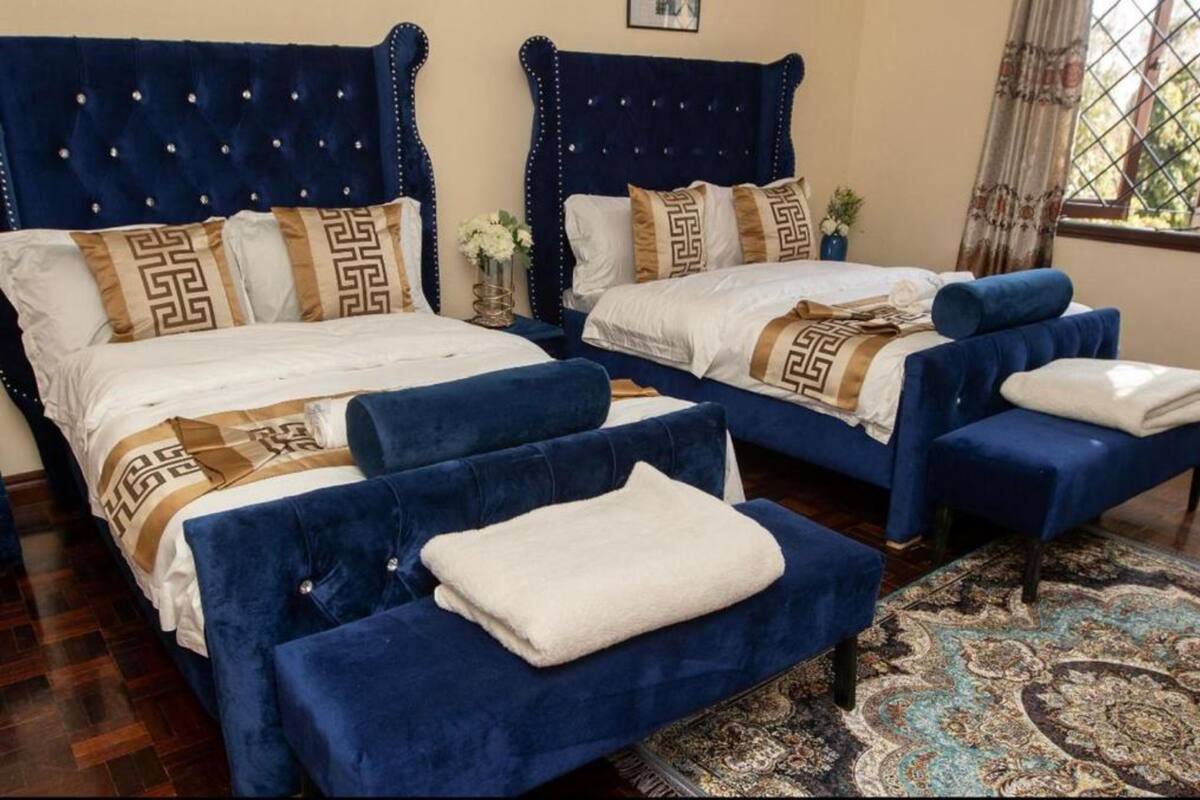 A hotel room with two beds, each with a blue headboard, white bedding, and decorative pillows. There are two blue armchairs and a small table with a white towel.
