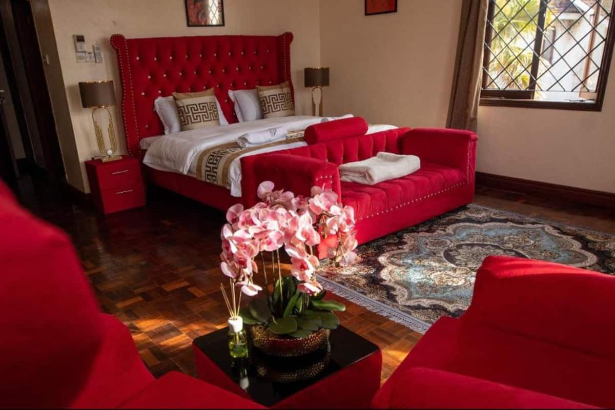 A bedroom with a red upholstered bed, a red sofa, a patterned rug, and a vase of pink orchids on a black table.