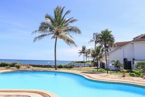 A swimming pool with a curved edge, surrounded by palm trees and a building with a tiled roof.