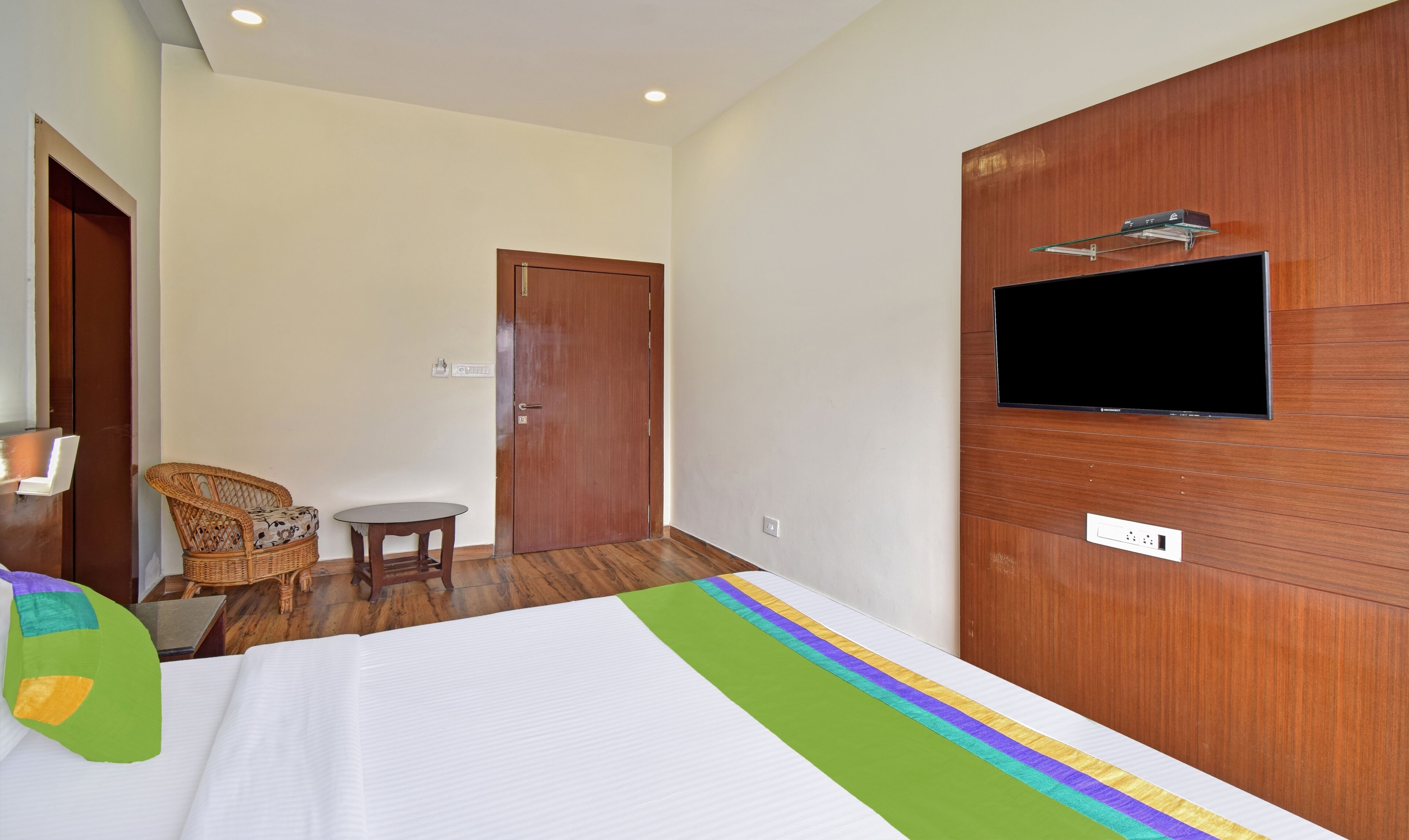 A hotel room with a bed, a television mounted on the wall, a small table, a wicker chair, and a door.