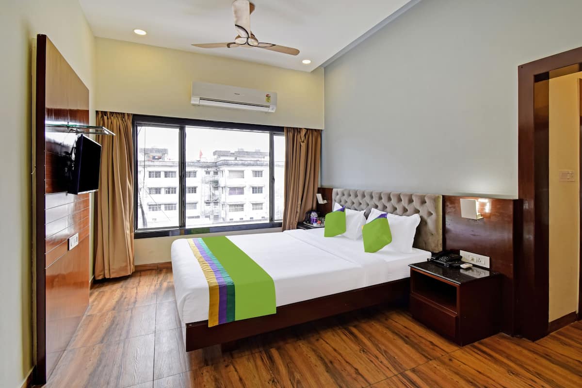 A hotel room with a bed, a TV, an air conditioning unit, a bedside table, and a window with a view of buildings.