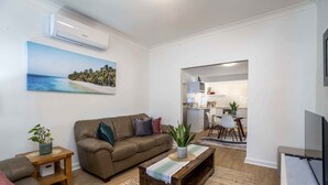 A living room with a brown sofa, a coffee table, and a TV. There is a painting of a beach on the wall and a kitchen in the background. - Bright, Cosy and Central Cottage- Free Wi-fi, Netflix and Disney Plus (Orange)