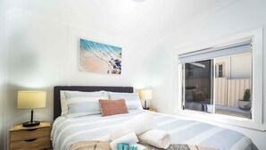 A bedroom with a bed, two bedside lamps, a nightstand, a window with a view of a house, and a beach-themed painting on the wall. - Bright, Cosy and Central Cottage- Free Wi-fi, Netflix and Disney Plus (Orange)