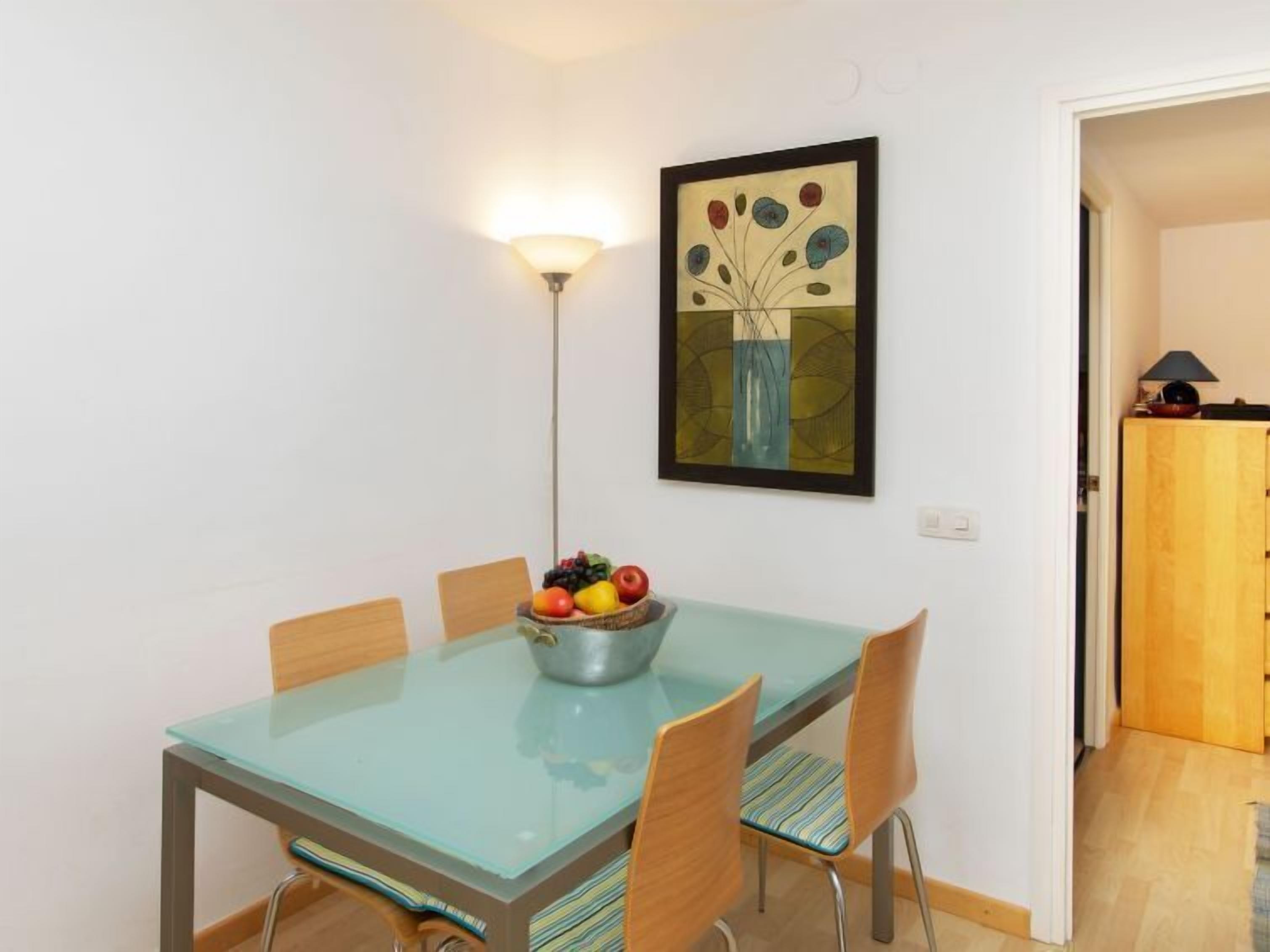 Un comedor con una mesa de vidrio, cuatro sillas, un frutero y una lámpara de pared.