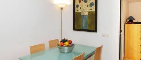 Un comedor con una mesa de vidrio, cuatro sillas, un frutero y una lámpara de pared.