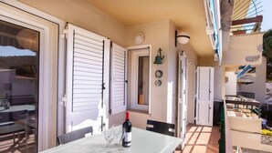 Una mesa de vidrio con una botella y copas, contraventanas blancas y vista a un edificio.