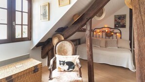 A cozy bedroom with a sloped ceiling, a bed with patterned pillows, a chair with a cat pillow, and a rustic wooden beam.