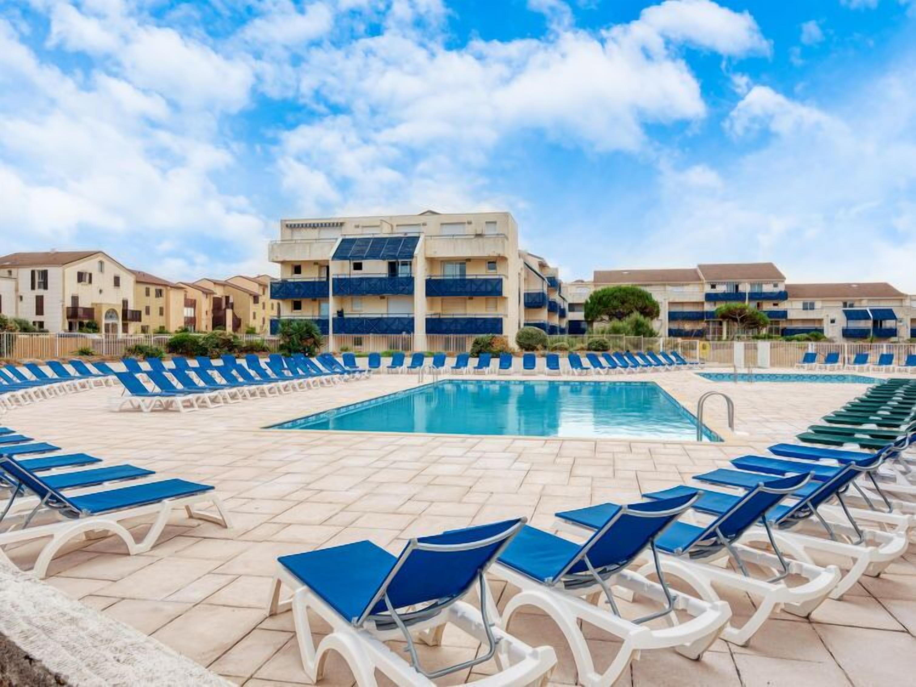 Una piscina con sillas reclinables azules dispuestas a su alrededor, rodeada de edificios de apartamentos con balcones.