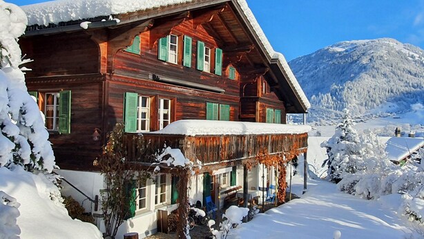 A wooden chalet with green shutters, a balcony, and a snowy mountain backdrop. - Hübeli (Kopp) by Interhome (Zweisimmen)