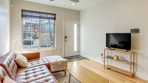 A living room with a leather sofa, a wooden coffee table, a TV on a wooden stand, and a large window with blinds. - 3-Story Philly Townhouse 2 Mi to University City! (Philadelphia)