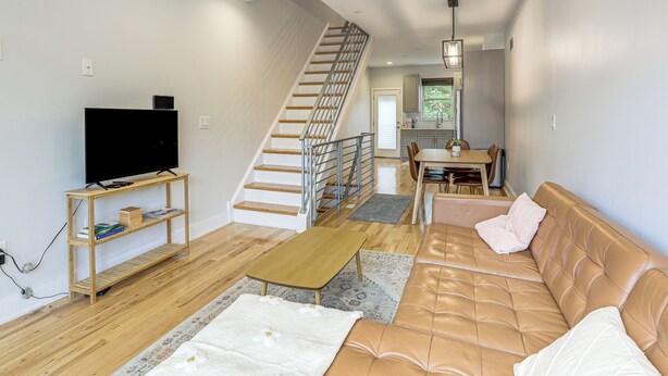 A living room with a staircase, a sofa, a television on a shelf, and a dining area. - 3-Story Philly Townhouse 2 Mi to University City! (Philadelphia)