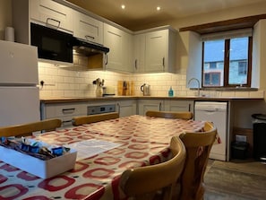 Uma cozinha compacta com armários brancos, forno embutido e máquina de lavar louça. Uma mesa de jantar com toalha de mesa estampada está posta com cadeiras.