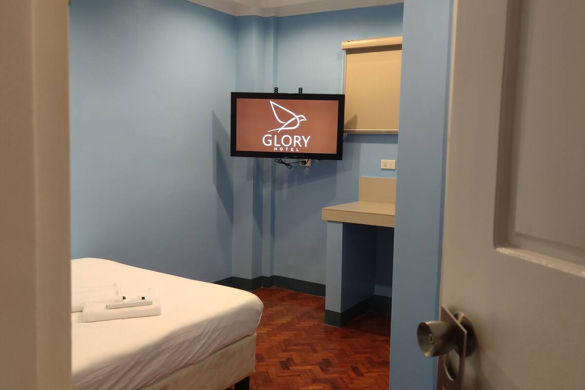 Une chambre d’hôtel avec un lit, un téléviseur affichant le nom de l’hôtel, une petite salle de bain et un plancher en bois.