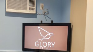 A hotel room with a mounted television displaying the hotel's name, a potted plant, a small wooden table with a phone and remote, and a doorbell with a sign. - Glory Hotel Cubao (Quezon City)