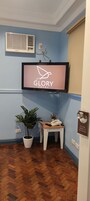 A hotel room with a mounted television displaying the hotel's name, a potted plant, a small wooden table with a phone and remote, and a doorbell with a sign.