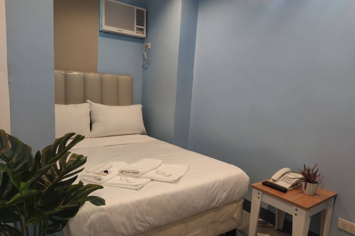 Un lit bien fait, avec du linge de lit blanc, une table de chevet avec un téléphone et une plante, et un climatiseur fixé au mur.