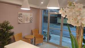 A reception area with a white reception desk, two wicker chairs, a small round table, a potted plant, and a large window offering a view of the street. - Glory Hotel Cubao (Quezon City)