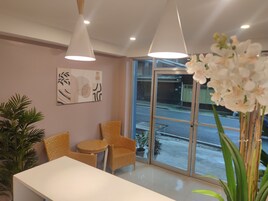 A reception area with a white reception desk, two wicker chairs, a small round table, a potted plant, and a large window offering a view of the street.