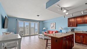 A spacious living area with a large sliding glass door leading to a balcony, a kitchen with wooden cabinets, and a dining area with a white table and chairs. - Laketown Wharf 913 (Panama City Beach)