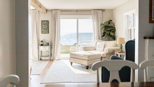 A cozy living room with a view of the sea, a comfortable sofa, a small table with a lamp, and a large plant. - Bayside Bungalow: Autumn Lakefront Escape | 4BR, Firepit & Quiet Surroundings (Hessel)